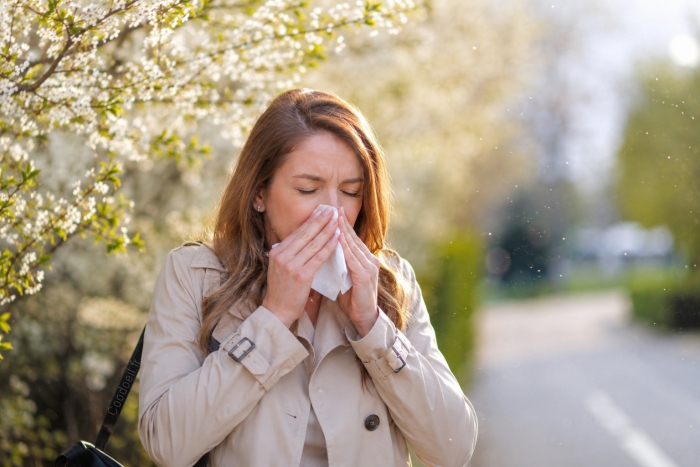 Une femme &eacute;ternue dans un mouchoir au milieu d&rsquo;arbres en fleurs lors d&rsquo;une journ&eacute;e de printemps, illustrant les allergies saisonni&egrave;res provoqu&eacute;es par le pollen.